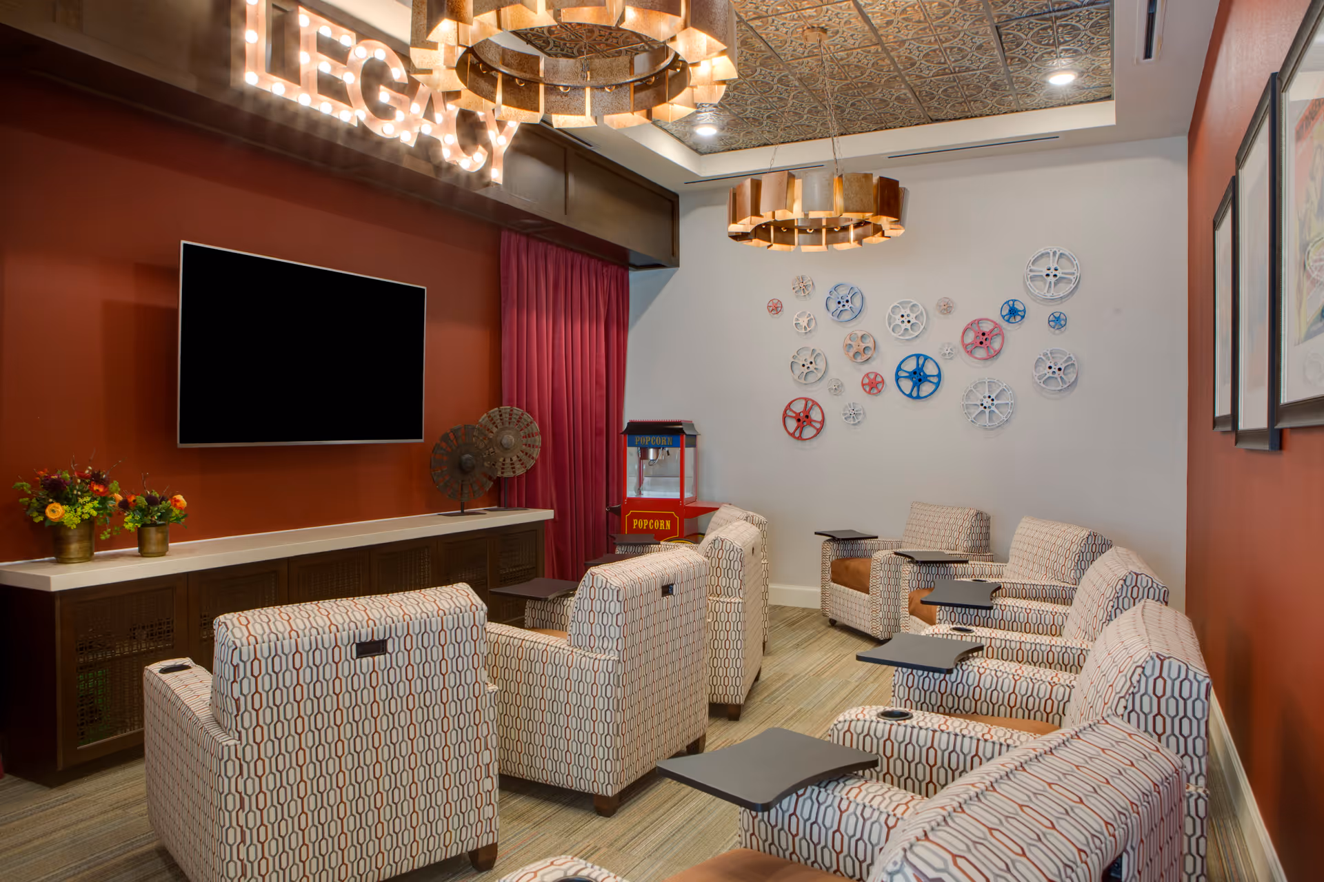 A cozy media room with patterned armchairs arranged in rows facing a large flat-screen TV mounted on a red accent wall. Above the TV, illuminated letters spell out 'LEGACY'. The room features decorative ceiling lights, a popcorn machine in the corner, and a wall adorned with colorful film reel decorations. There are also framed pictures on the adjacent wall and a cabinet with flower arrangements below the TV.