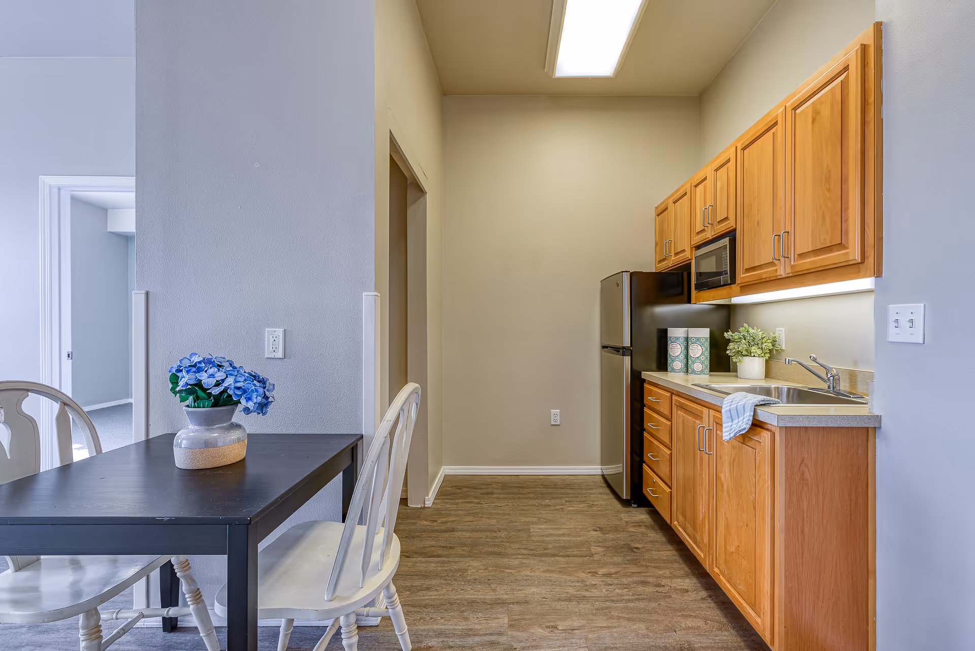 Small kitchen and dining area with wooden cabinets, a stainless refrigerator and sink, and a table with chairs and a vase of blue flowers.
