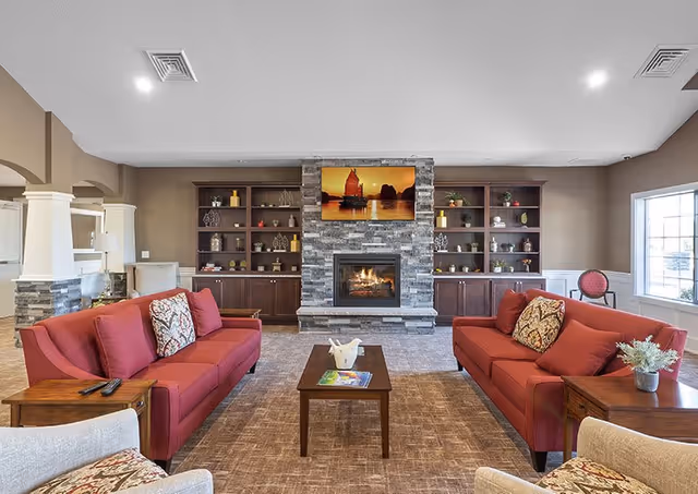 A cozy living room with two red sofas facing each other across a wooden coffee table. The room features a stone fireplace with a flat-screen TV mounted above it, flanked by built-in wooden shelves decorated with various ornaments. There are large windows allowing natural light to fill the space, and additional seating is visible in the foreground and background.