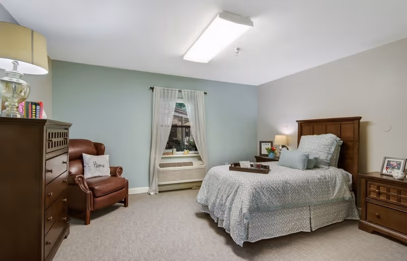A cozy bedroom with a single bed covered in a patterned light blue and white bedspread, a wooden headboard, and matching wooden nightstands on either side. One nightstand has a lamp and framed photos, while the other has a lamp and a small plant. There is a brown leather armchair with a 'Home' pillow next to a wooden dresser. A window with sheer white curtains is centered on the light blue accent wall, and a heating/cooling unit is below the window. The room has neutral-colored carpet and soft lighting.