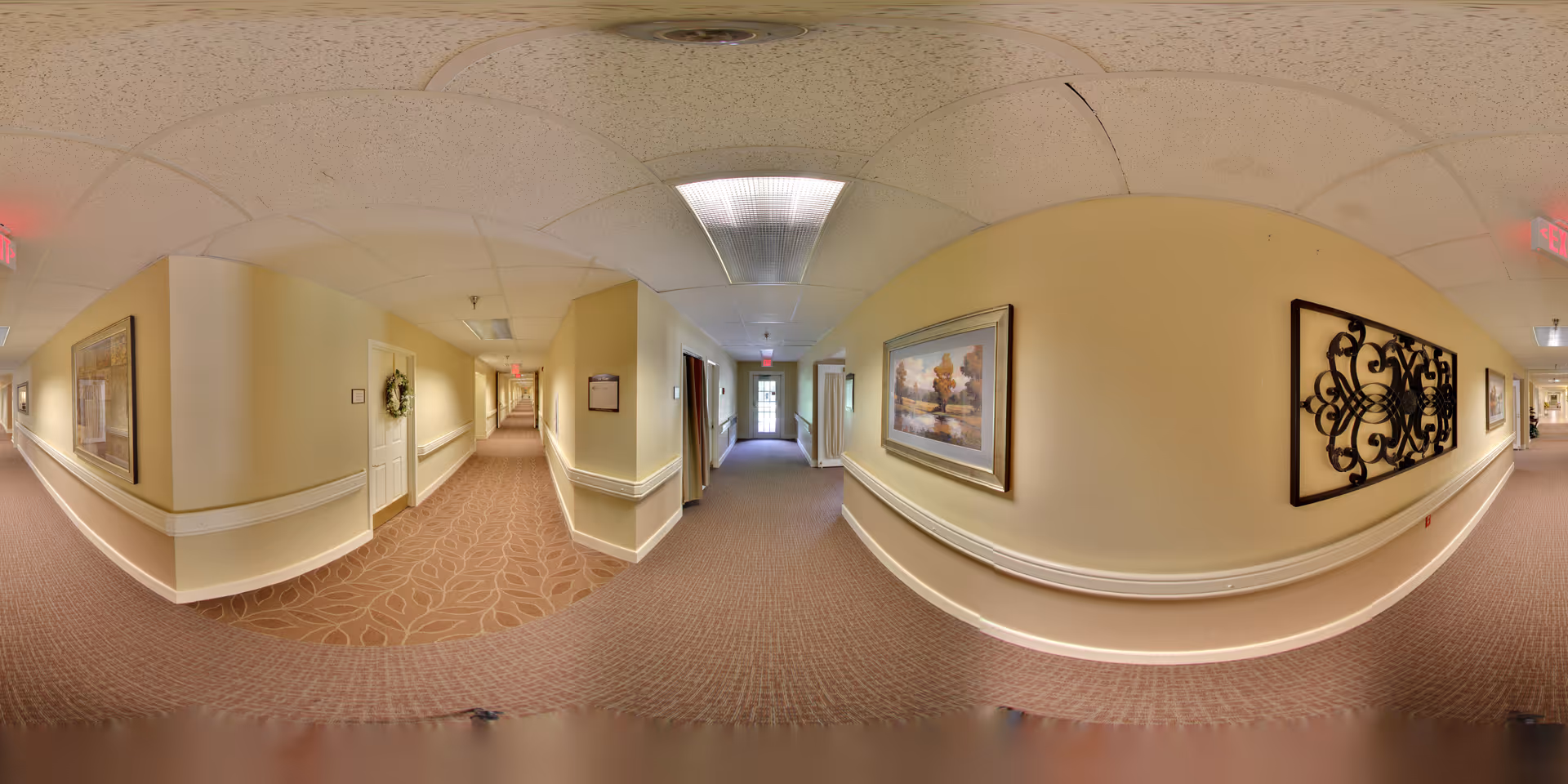A wide-angle view of a long, carpeted hallway in a senior living facility with beige walls and ceiling tiles. The hallway is decorated with framed artwork and a decorative metal wall piece. There are doors along the hallway, some with wreaths, and handrails on both sides. Exit signs are visible at both ends of the hallway.