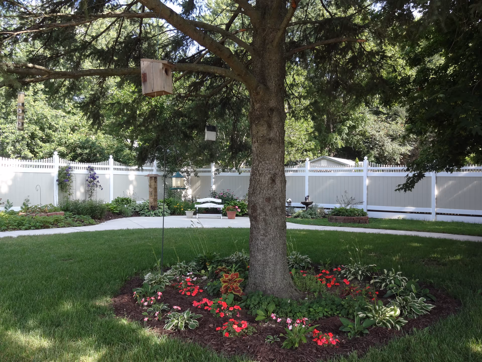 A peaceful outdoor garden area with a large tree in the center surrounded by colorful flowers and plants. There is a white fence enclosing the garden, a white bench, bird feeders hanging from the tree, and a paved walkway curving through the green lawn.