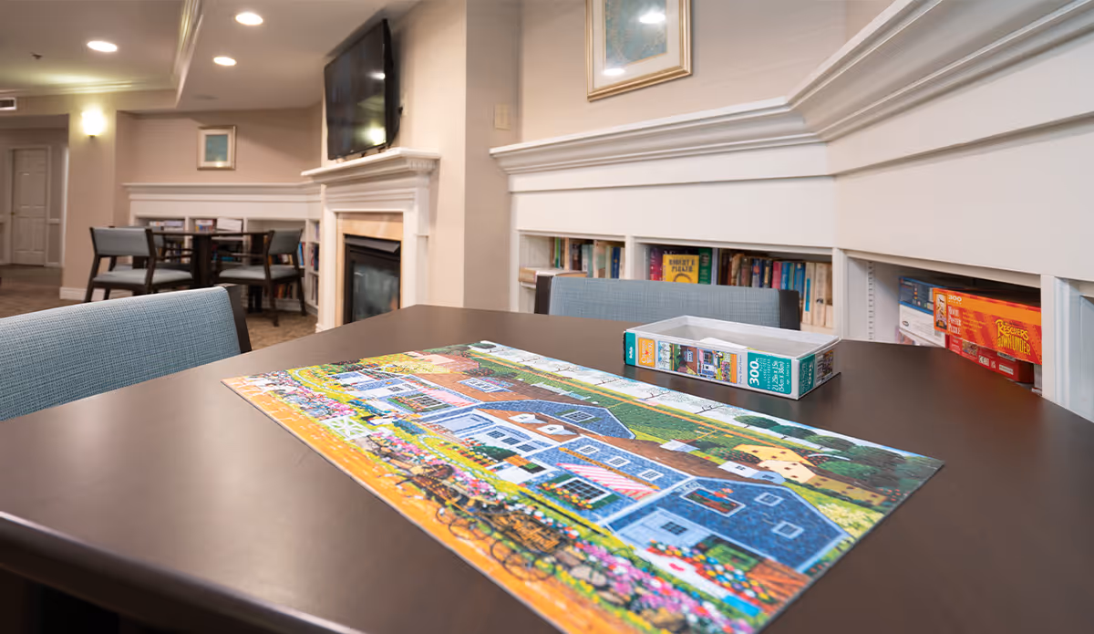A cozy common area with a dark wooden table featuring a partially completed colorful jigsaw puzzle. Surrounding the table are light blue upholstered chairs. In the background, there is a fireplace with a mounted flat-screen TV above it, built-in bookshelves filled with books and games, and additional tables and chairs. The room has soft lighting and neutral-colored walls.