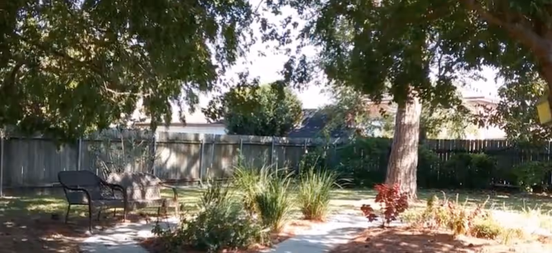 A peaceful outdoor garden area with a paved walkway, surrounded by green plants and trees providing shade. There is a black metal bench on the left side under the trees, and a wooden fence enclosing the space in the background.