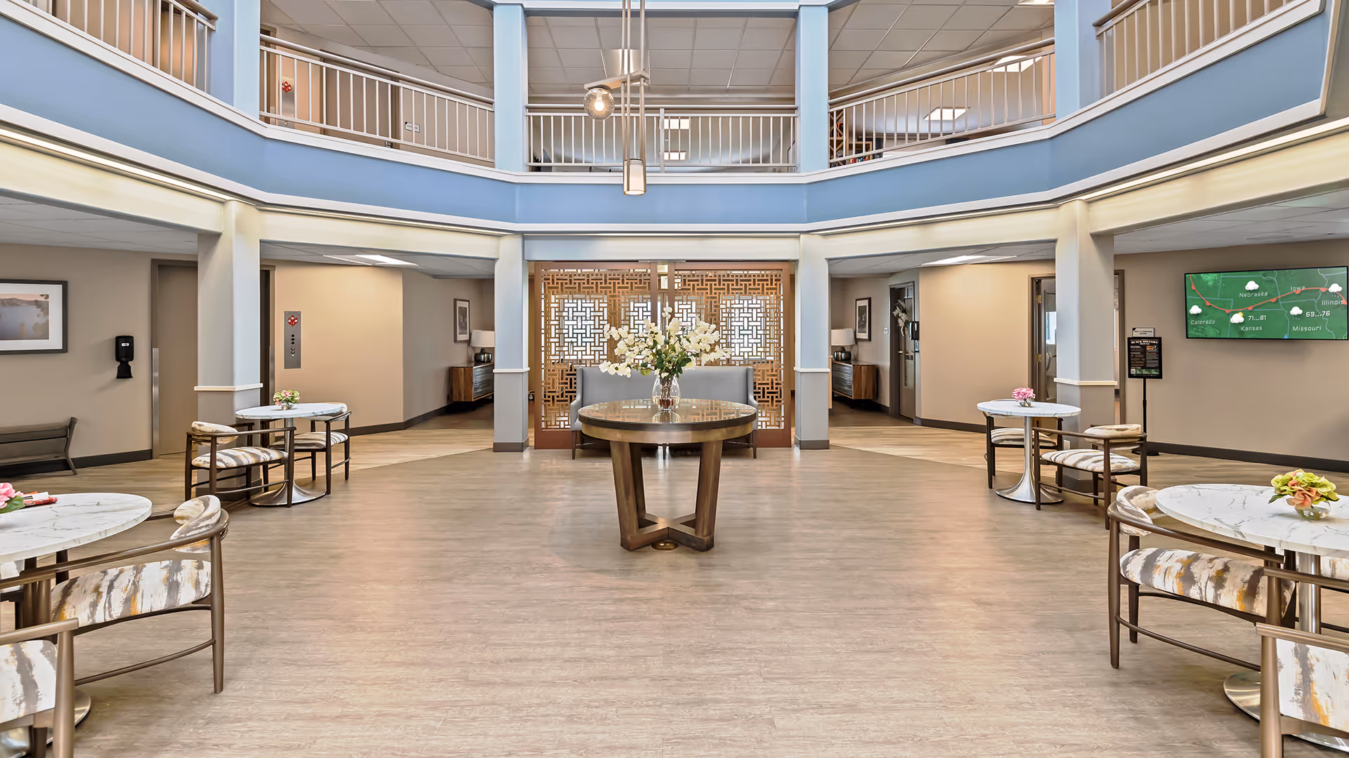 Spacious and well-lit common area in a senior living facility with a round wooden table in the center holding a vase of white flowers. Surrounding the area are small round tables with chairs, each table decorated with small flower arrangements. The room features a high ceiling with a second-floor balcony railing, beige walls, and a decorative wooden partition in the background. A television screen displaying weather information is mounted on the right wall.