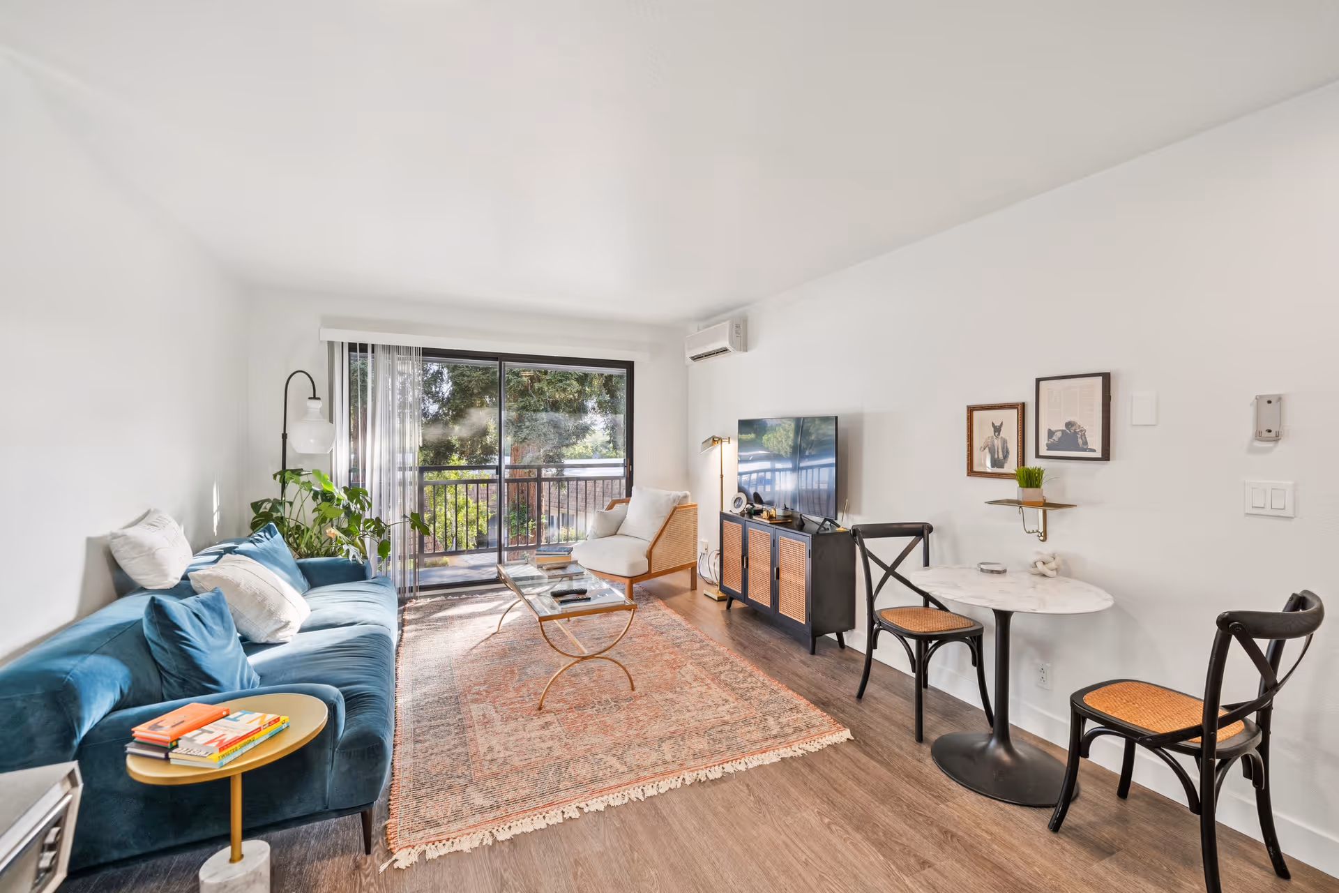 Bright living room with a blue sofa, a small round side table with books, a glass coffee table on a patterned rug, a white armchair near sliding glass doors leading to a balcony with trees outside, a TV on a wooden console, and a small round dining table with two chairs against a white wall with framed pictures and a small shelf with a plant.