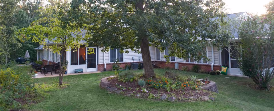 Single-story building with a grassy courtyard, a central tree surrounded by a flower bed, and a patio with outdoor seating.