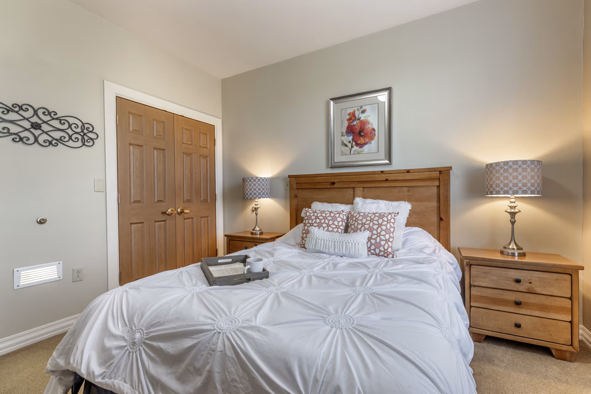 A cozy bedroom with a wooden bed frame and white bedding adorned with decorative pillows. There are two wooden nightstands on either side of the bed, each with a lamp featuring a patterned lampshade. A framed floral artwork hangs above the bed, and a double wooden closet door is visible on the left side of the room.