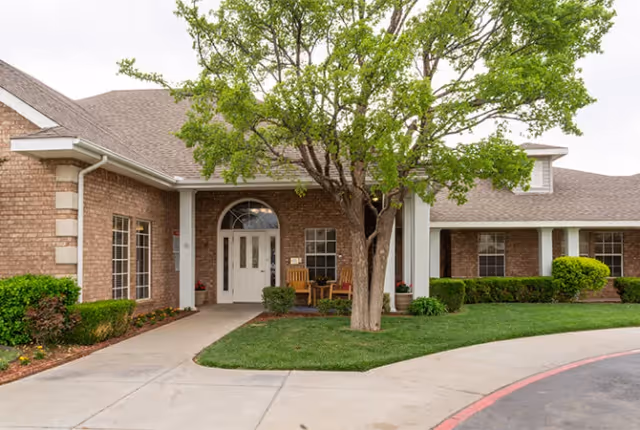 Exterior view of Plum Creek Place featuring a brick building with a covered entrance, white double doors, a tree in front, and neatly trimmed bushes and grass along the walkway.