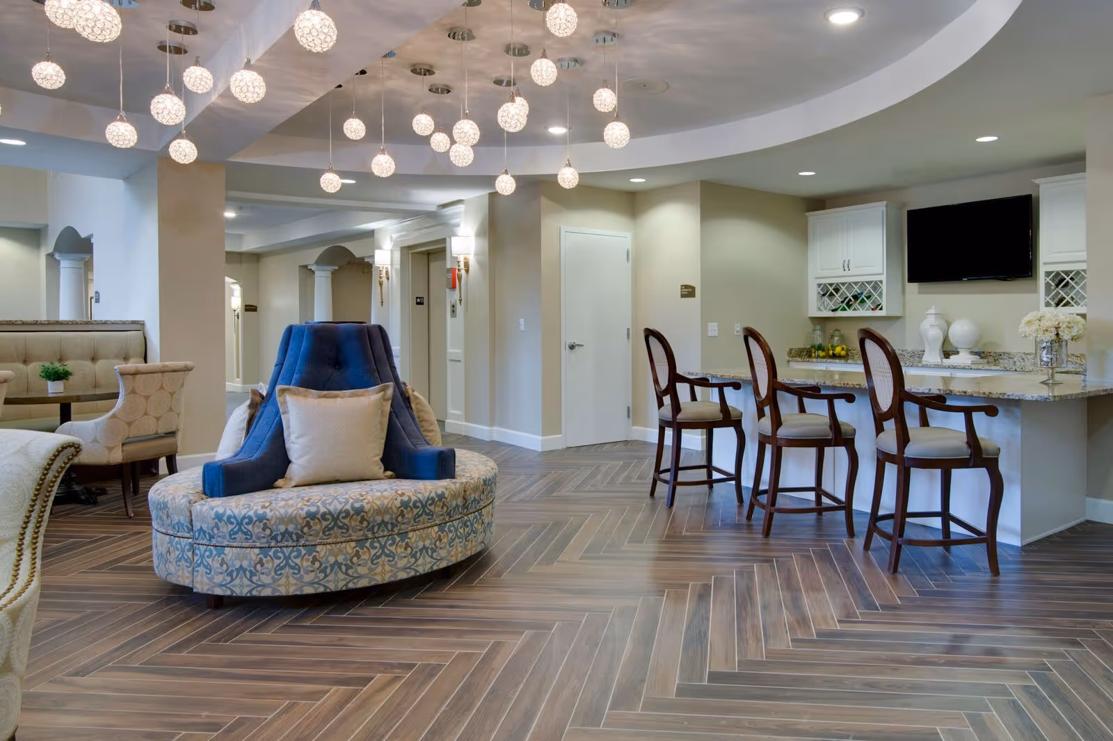 A spacious and elegant common area in a senior living facility featuring a round cushioned seating area with blue and patterned upholstery, a row of high wooden chairs at a granite countertop bar, beige walls, modern pendant lights hanging from the ceiling, and a flat-screen TV mounted on the wall.