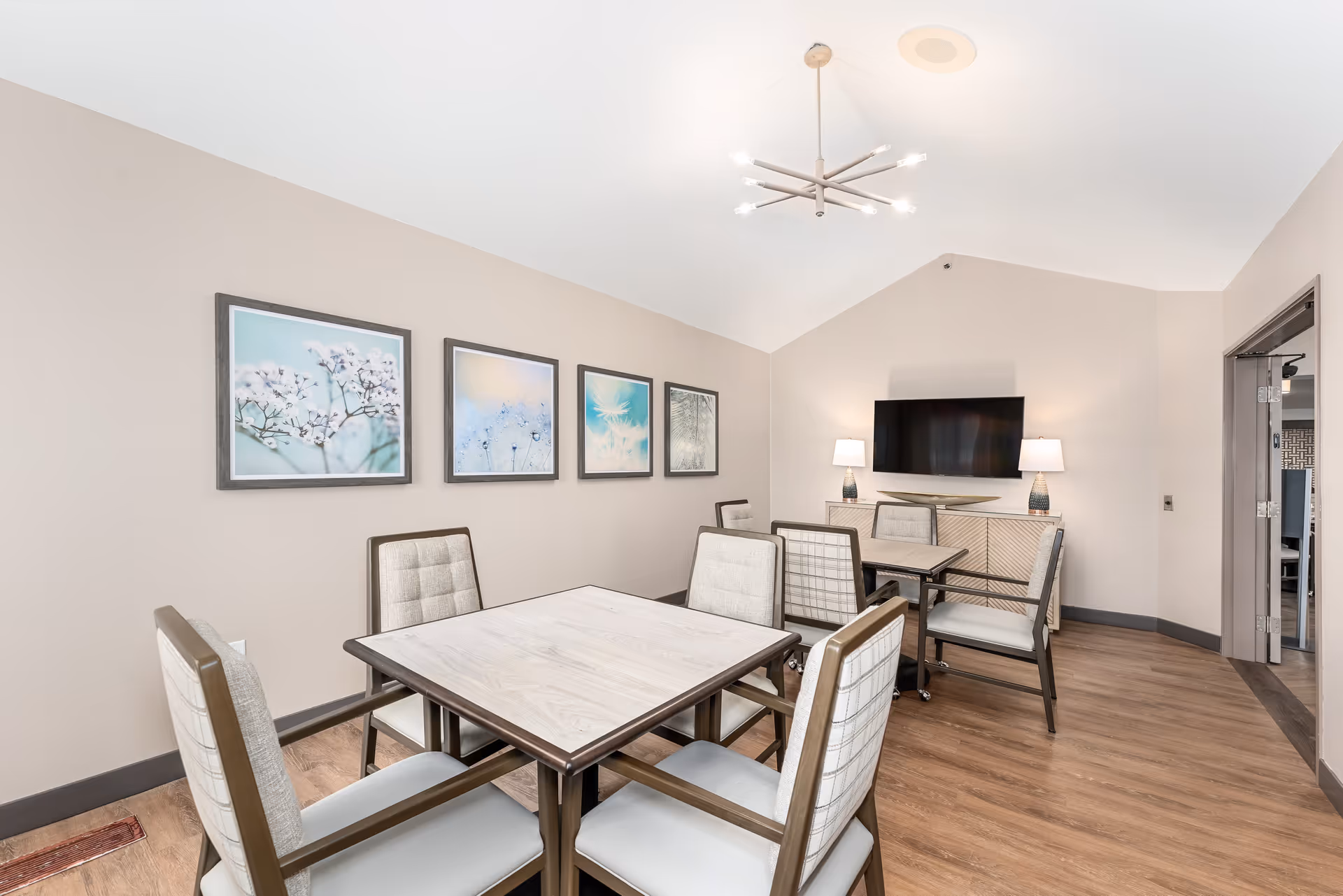 A bright and modern common area with two square tables, each surrounded by four cushioned chairs. The room has light beige walls adorned with four framed pictures of delicate floral and nature scenes. A flat-screen TV is mounted on the far wall above a sideboard with two table lamps. The floor is wood, and a contemporary chandelier hangs from the vaulted ceiling.