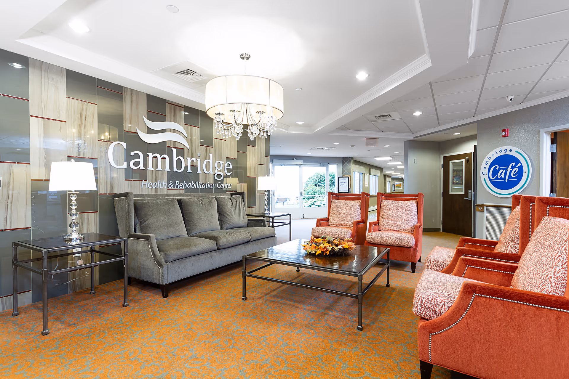 A bright and spacious lounge area in Cambridge Health and Rehabilitation Center featuring a gray sofa, four orange upholstered armchairs, a large coffee table with a floral centerpiece, two side tables with lamps, a chandelier, and a wall with the Cambridge Health & Rehabilitation Center logo. There is a sign for Cambridge Café on the wall near a doorway.