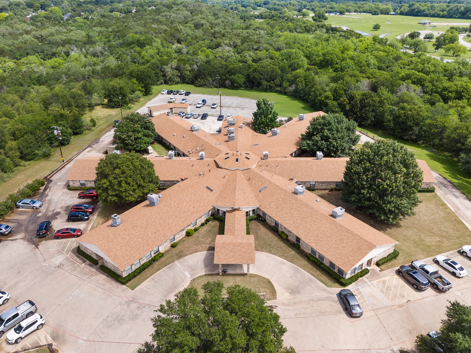 Aerial view of a star-shaped building with a brown roof surrounded by trees and parking lots with several cars parked. The building is located in a green, wooded area with open fields in the background.