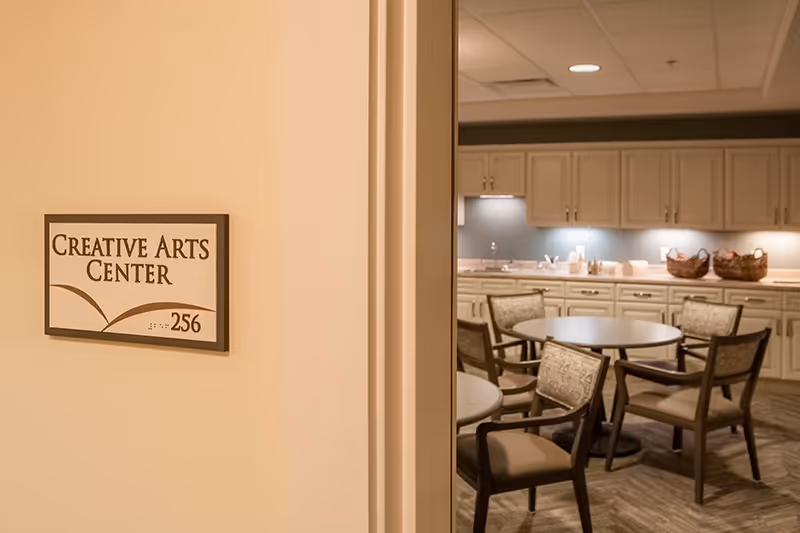 View of a room labeled Creative Arts Center with a sign on the wall showing room number 256. Inside the room, there are round tables with chairs and a counter with cabinets and baskets, suggesting a space for creative activities or crafts.