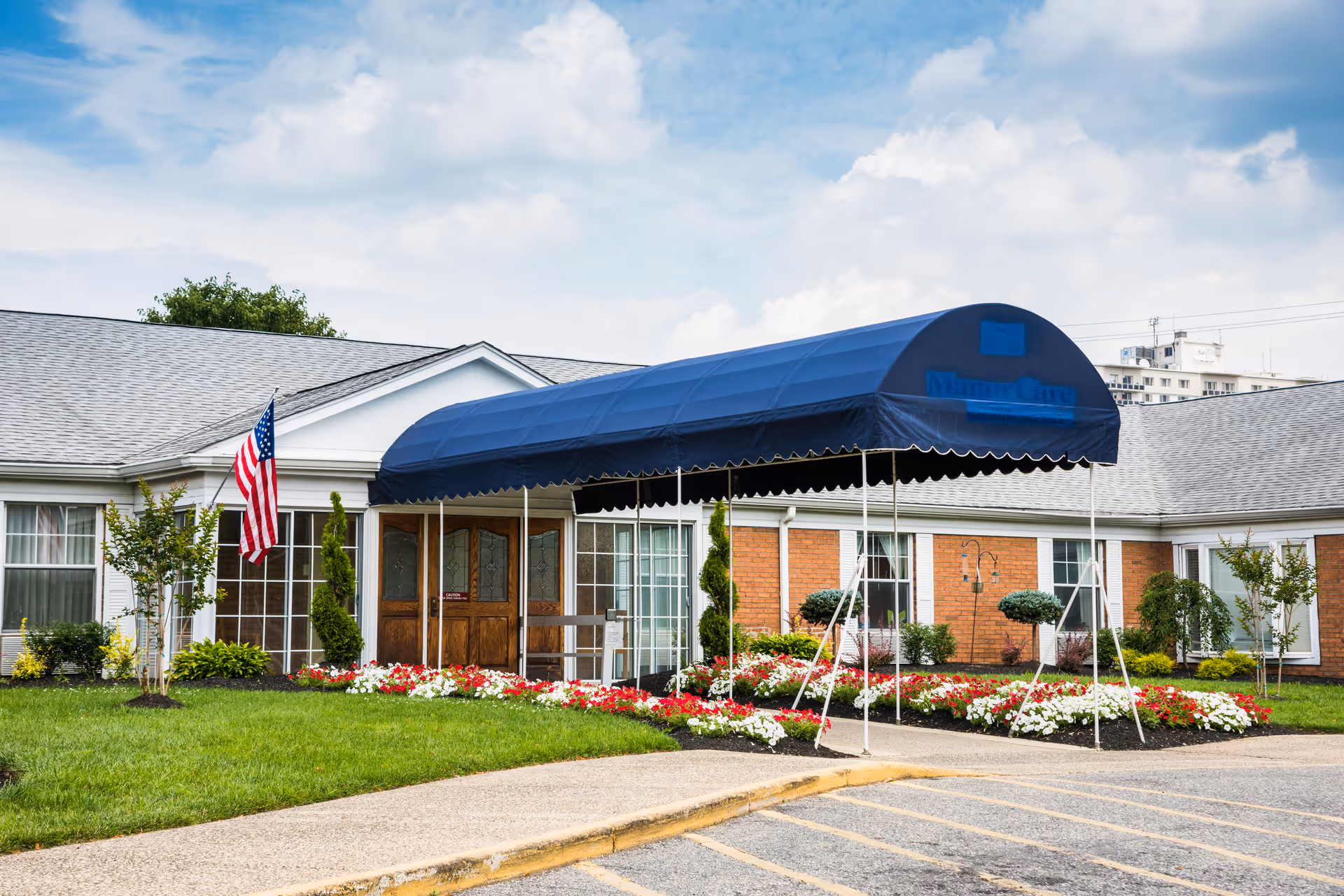 Exterior view of a single-story rehabilitation and health care center building with a blue canopy entrance, an American flag, well-maintained flower beds, and a paved driveway.