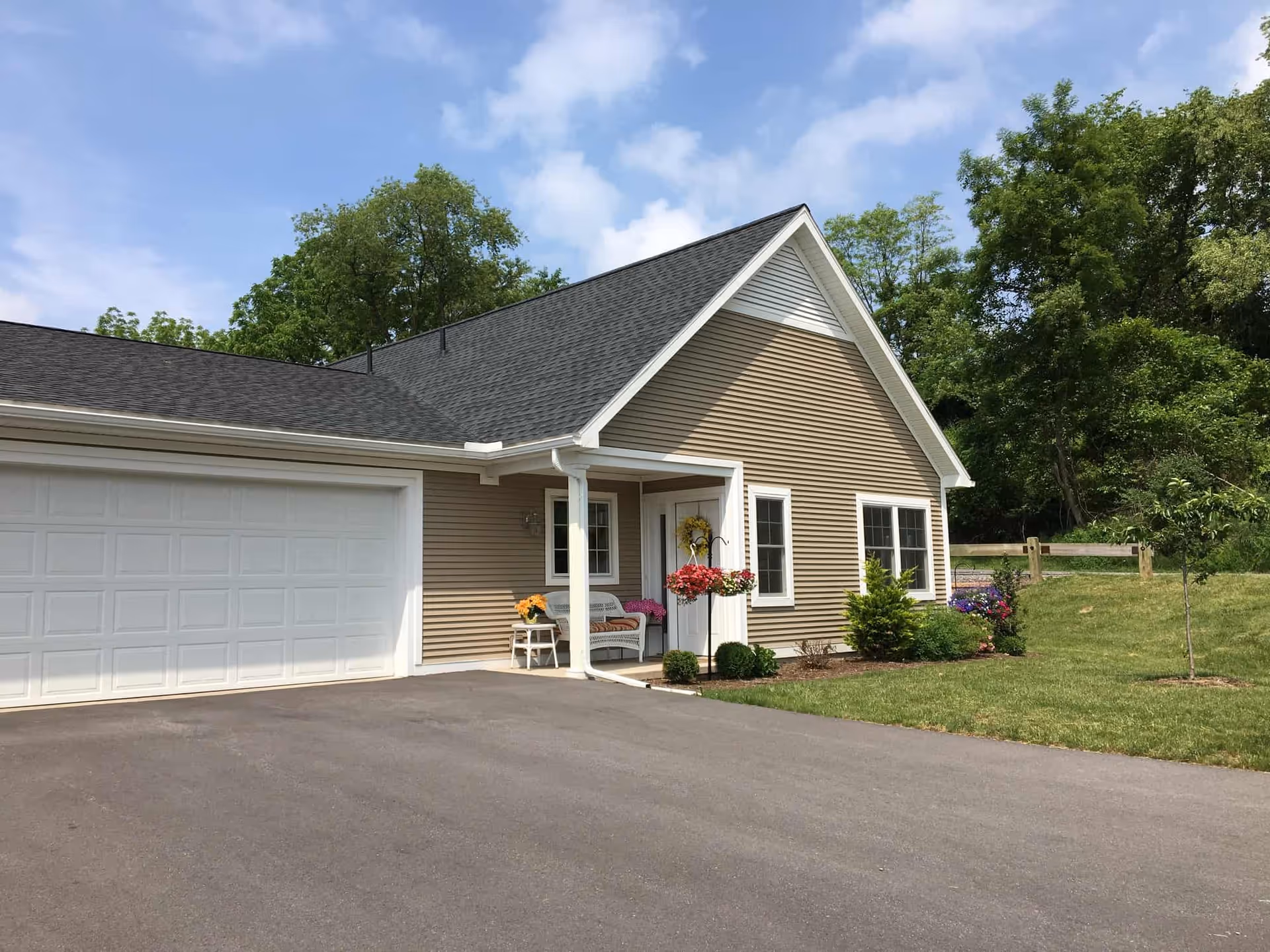 Exterior view of a single-story building with beige siding and a dark shingled roof. The building has a white garage door on the left and a white front door with a small covered porch. There is a white bench with cushions and a small table with a flower pot on the porch. The surrounding area includes a paved driveway, green grass, some bushes, flowers, and trees under a partly cloudy sky.