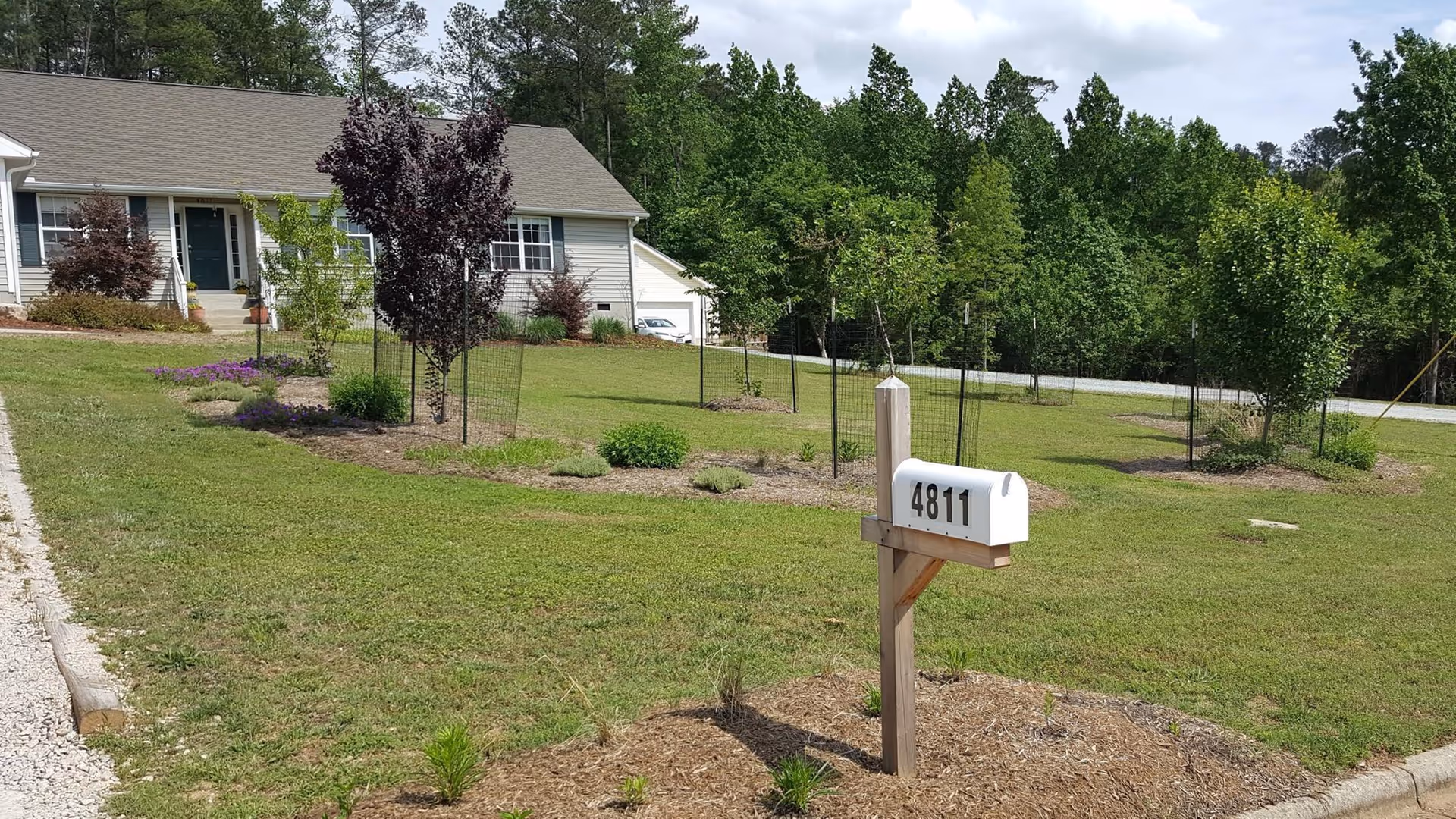 A single-story house with a beige exterior and a gray roof, surrounded by a well-maintained lawn with small trees and shrubs. A white mailbox with the number 4811 is mounted on a wooden post near the curb. The background features a wooded area with tall green trees under a partly cloudy sky.