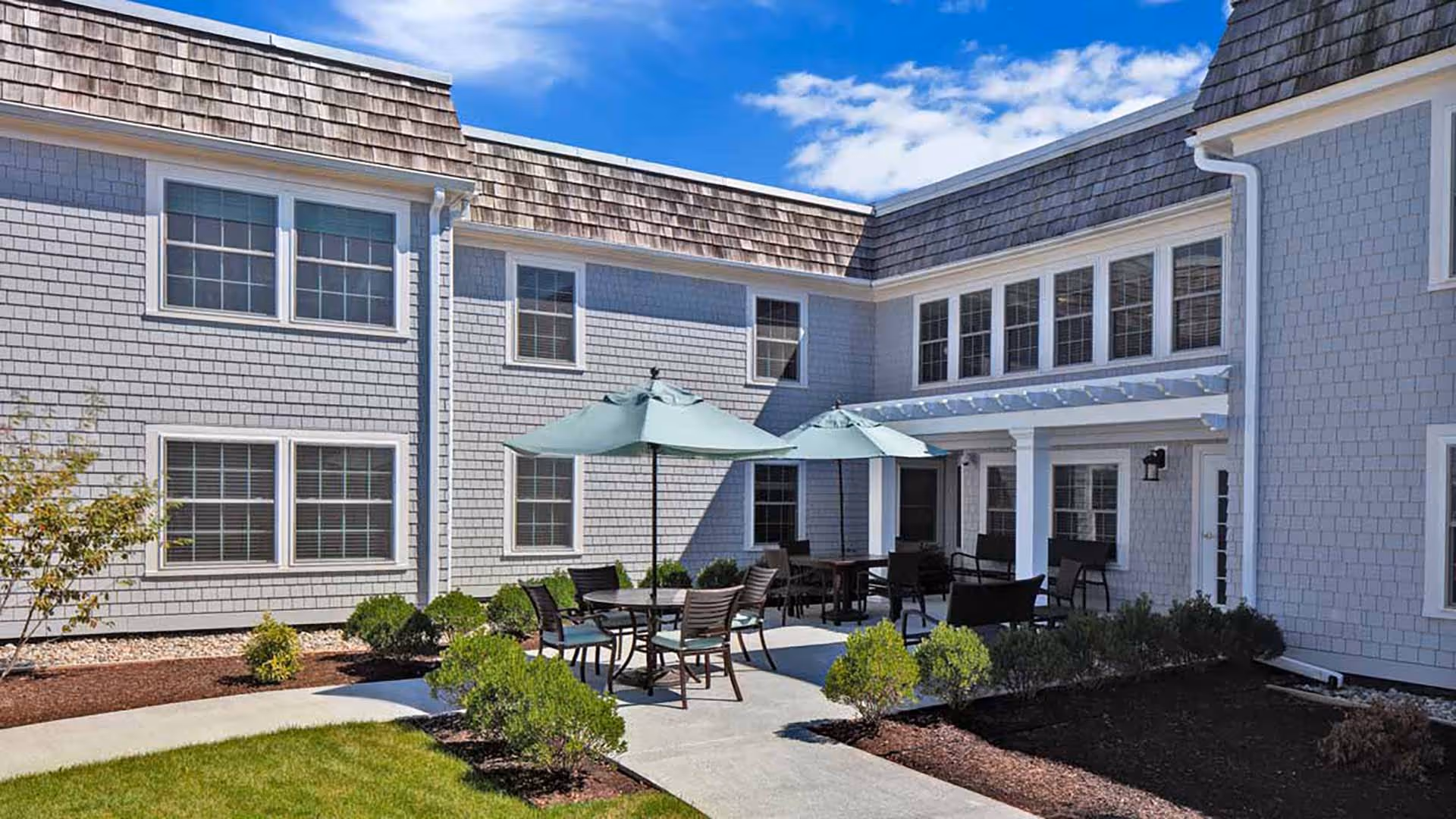 Outdoor courtyard area of a senior living facility with gray shingle siding and multiple windows. The courtyard features a concrete patio with round tables, chairs, and large green umbrellas. There are small bushes and landscaped garden beds surrounding the patio under a bright blue sky with some clouds.