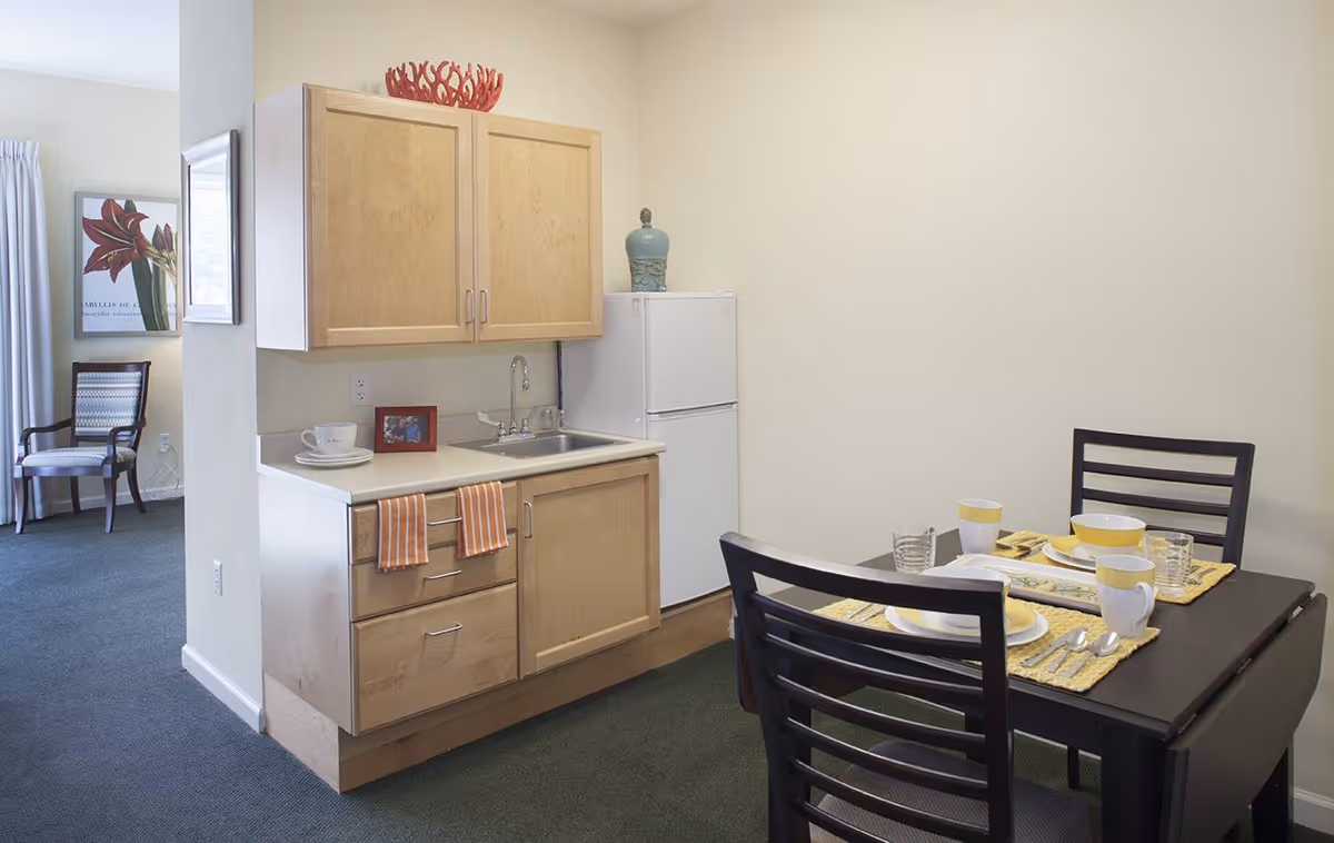Small kitchenette with light wood cabinets, a sink and mini fridge adjacent to a dining table set for two in an apartment.