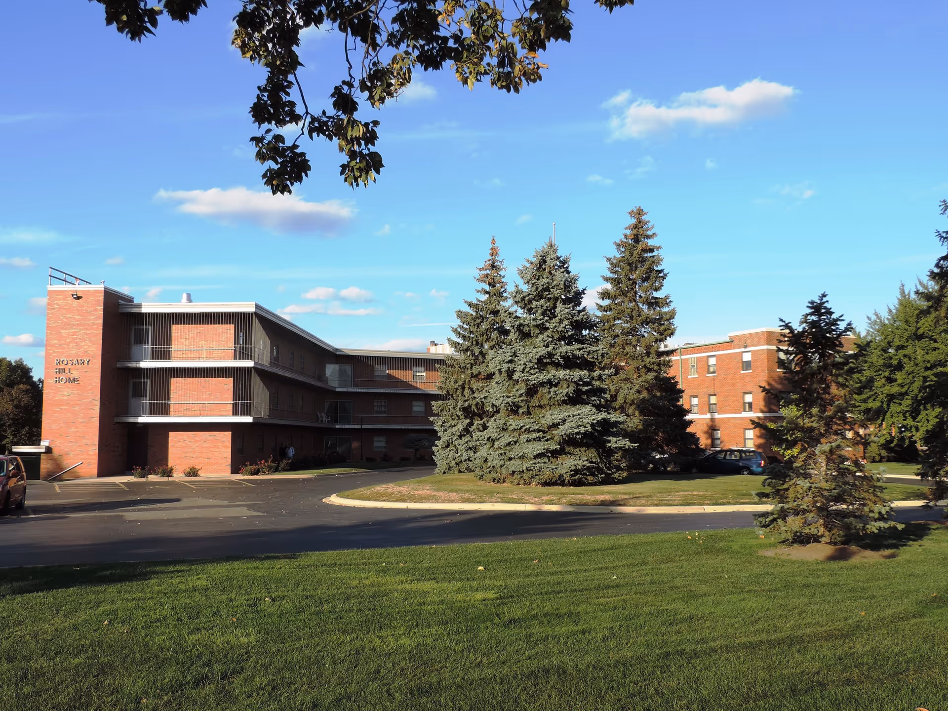 Exterior view of Rosary Hill Home, a multi-story brick building surrounded by green grass, several tall evergreen trees, and a clear blue sky with a few clouds.