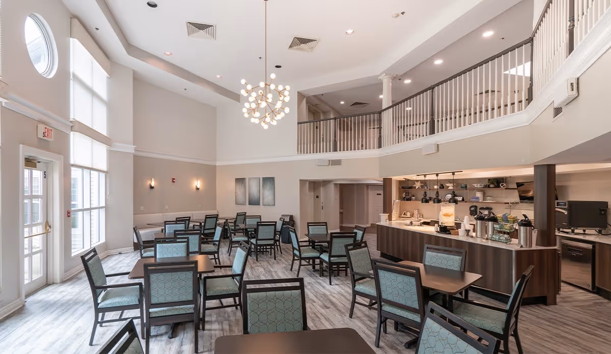 A spacious dining area with multiple tables and chairs featuring light blue patterned upholstery. The room has high ceilings with a modern chandelier, large windows allowing natural light, and a serving counter with coffee and beverage dispensers. There is a second-floor balcony overlooking the dining space.