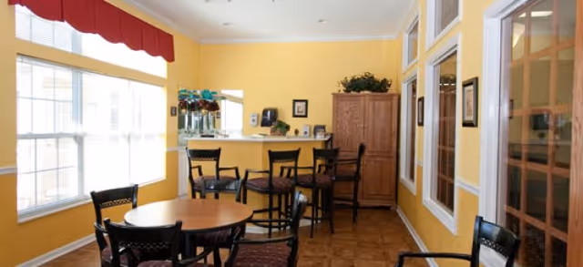 A bright room with yellow walls featuring a round wooden table surrounded by four chairs in the foreground. In the background, there is a counter with three high chairs, a wooden cabinet, and a large window with a red valance letting in natural light. The room has a warm and inviting atmosphere.