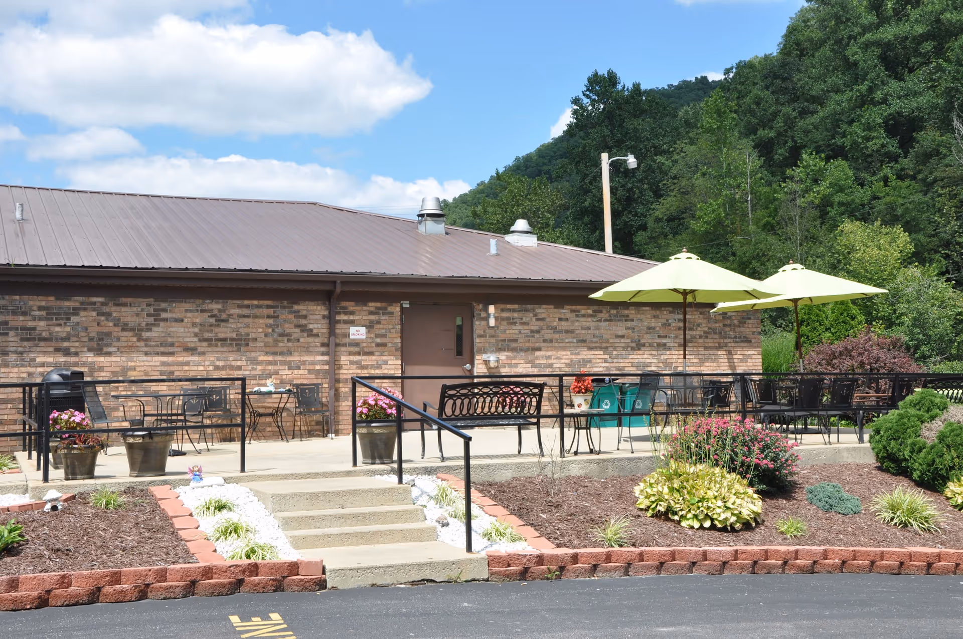 Outdoor patio area with tables, chairs, and umbrellas in front of a brick building. There are flower beds with various plants and shrubs surrounding the patio, and a set of concrete steps with a handrail leads up to the patio. Trees and a blue sky with clouds are visible in the background.