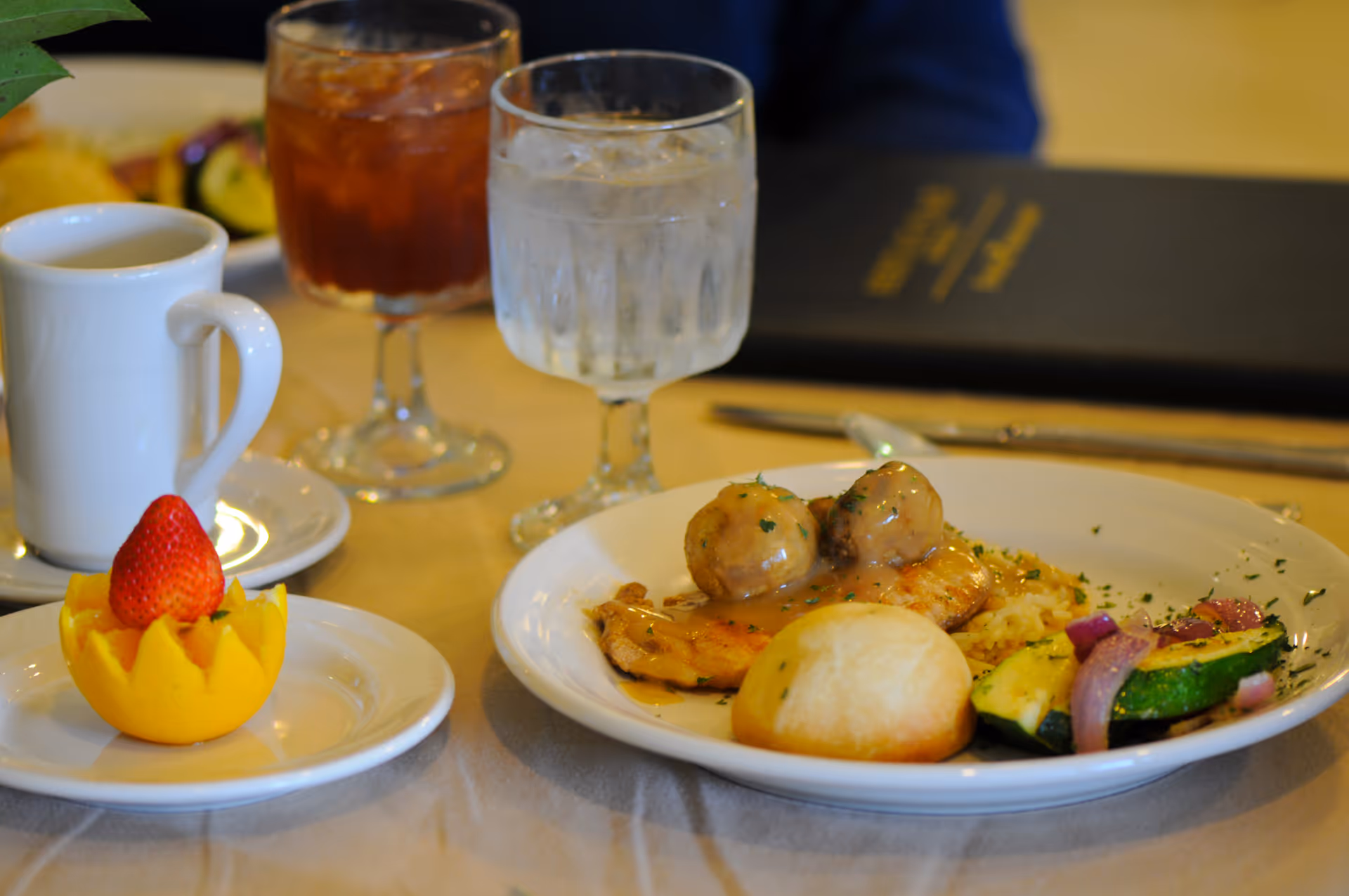 A dining table set with a plate of food including chicken with gravy and meatballs, rice, a bread roll, and sautéed vegetables. There is also a small dessert made of a strawberry placed in a hollowed-out orange peel, a cup of coffee, a glass of iced tea, and a glass of water. A menu is visible in the background.