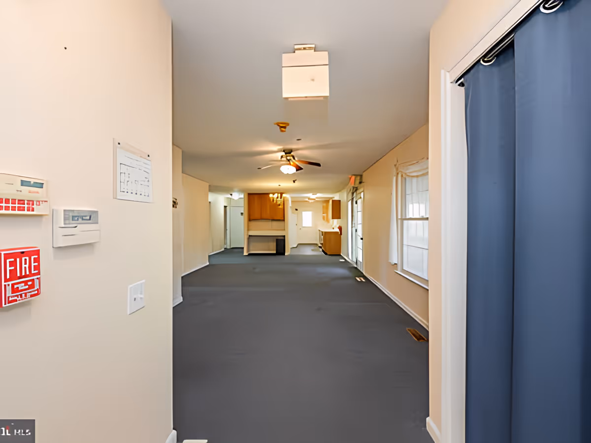 Long empty interior hallway/common area with carpet, ceiling fans, windows on the right and a kitchenette at the far end.