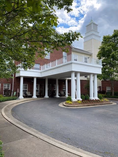 Exterior view of Belmont Village Senior Living Memphis showing a red brick building with white columns supporting a covered entrance. The building has multiple windows and a tower-like structure on the roof. Trees and landscaping surround the driveway leading to the entrance under a partly cloudy sky.