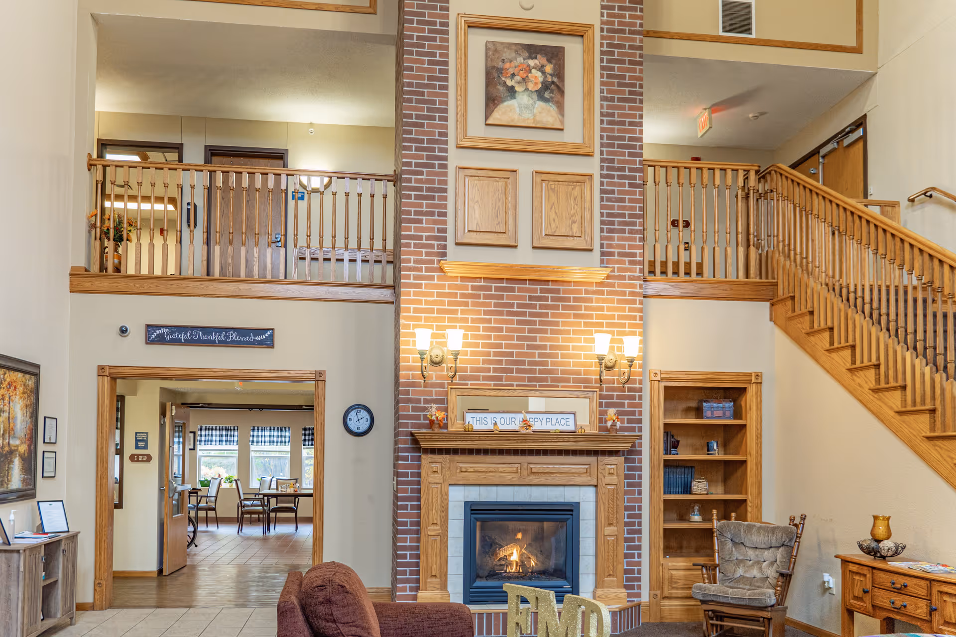 Interior view of a senior living facility lobby with a brick fireplace in the center, flanked by wooden bookshelves and a staircase on the right. There is a seating area with chairs and a couch in front of the fireplace. Above the fireplace is a framed floral painting and a sign that reads 'THIS IS OUR HAPPY PLACE'. The upper level has a wooden railing and doors leading to other rooms. The walls are painted beige with wooden trim.