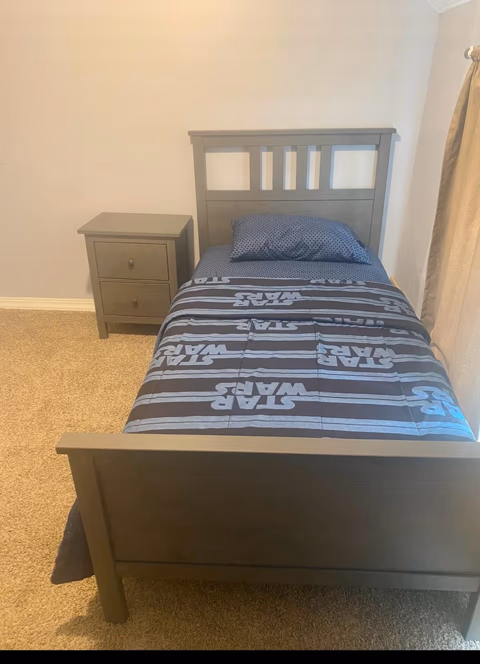 A small bedroom with a single gray wooden bed frame and matching nightstand. The bed is made with a Star Wars themed comforter and a blue pillow with a subtle pattern. The room has beige carpet and light-colored walls, with a curtain partially visible on the right side.