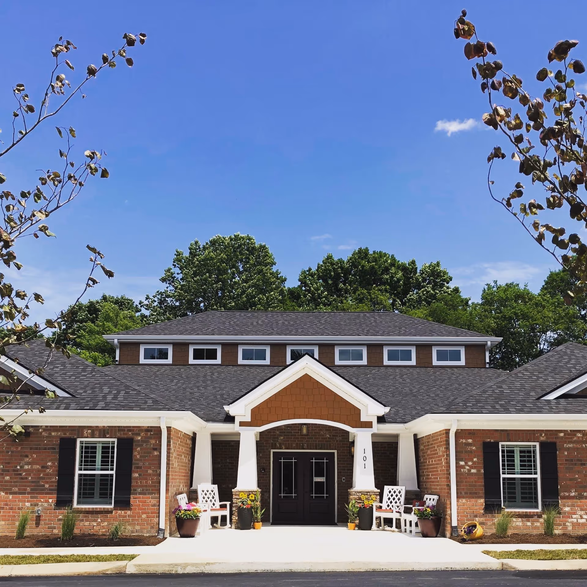Front exterior view of a single-story brick building with a dark shingled roof, white trim, and a covered entrance supported by white columns. There are two white benches and several flower pots with colorful flowers near the entrance. Trees and a clear blue sky are visible in the background.