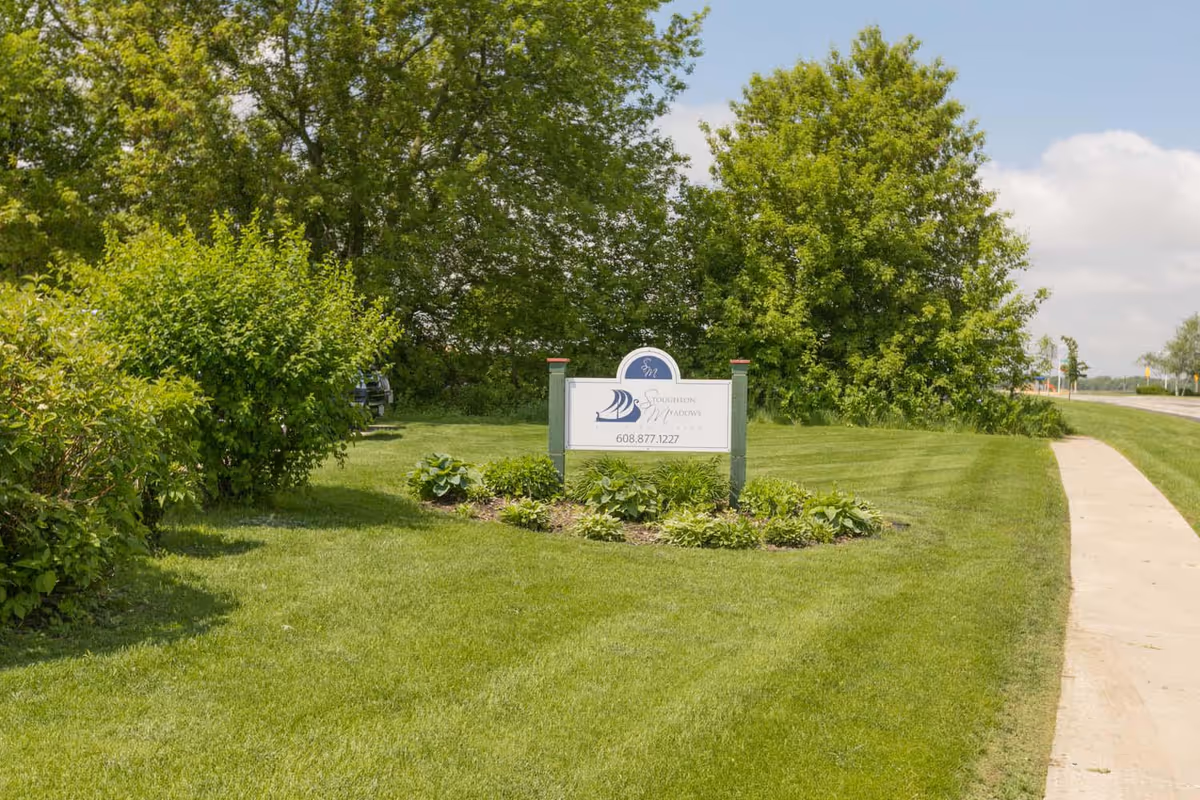 A landscaped lawn and sidewalk featuring a Stoughton Meadows Assisted Living sign surrounded by shrubs and trees.