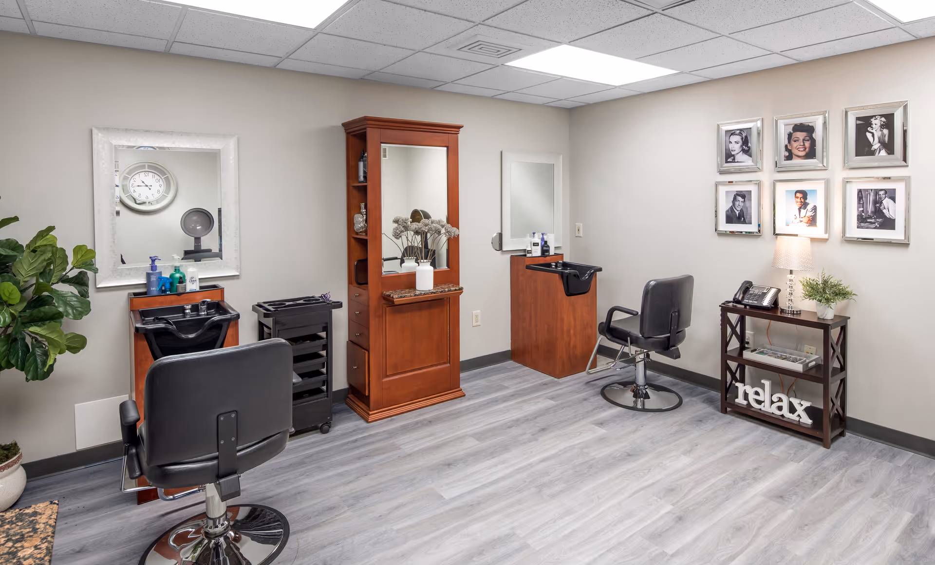Interior view of a hair salon area in a retirement community with two black salon chairs facing mirrors. There are wooden cabinets and a small table with a lamp, phone, plant, and decorative 'relax' sign. The walls are decorated with framed black and white photos of classic celebrities. The floor is light gray wood, and there is a potted plant in the corner.