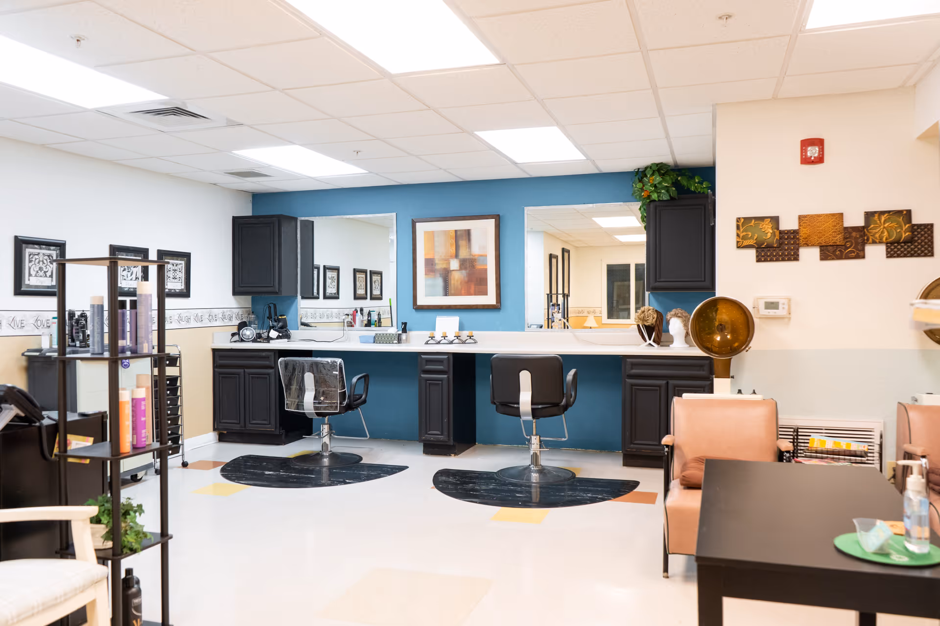 Interior view of a salon area in a senior living facility with two black salon chairs in front of a long counter with mirrors, black cabinets, and a blue accent wall. There are decorative items on the counter and wall, a hair dryer chair, and a black table with a green tray and hand sanitizer in the foreground.