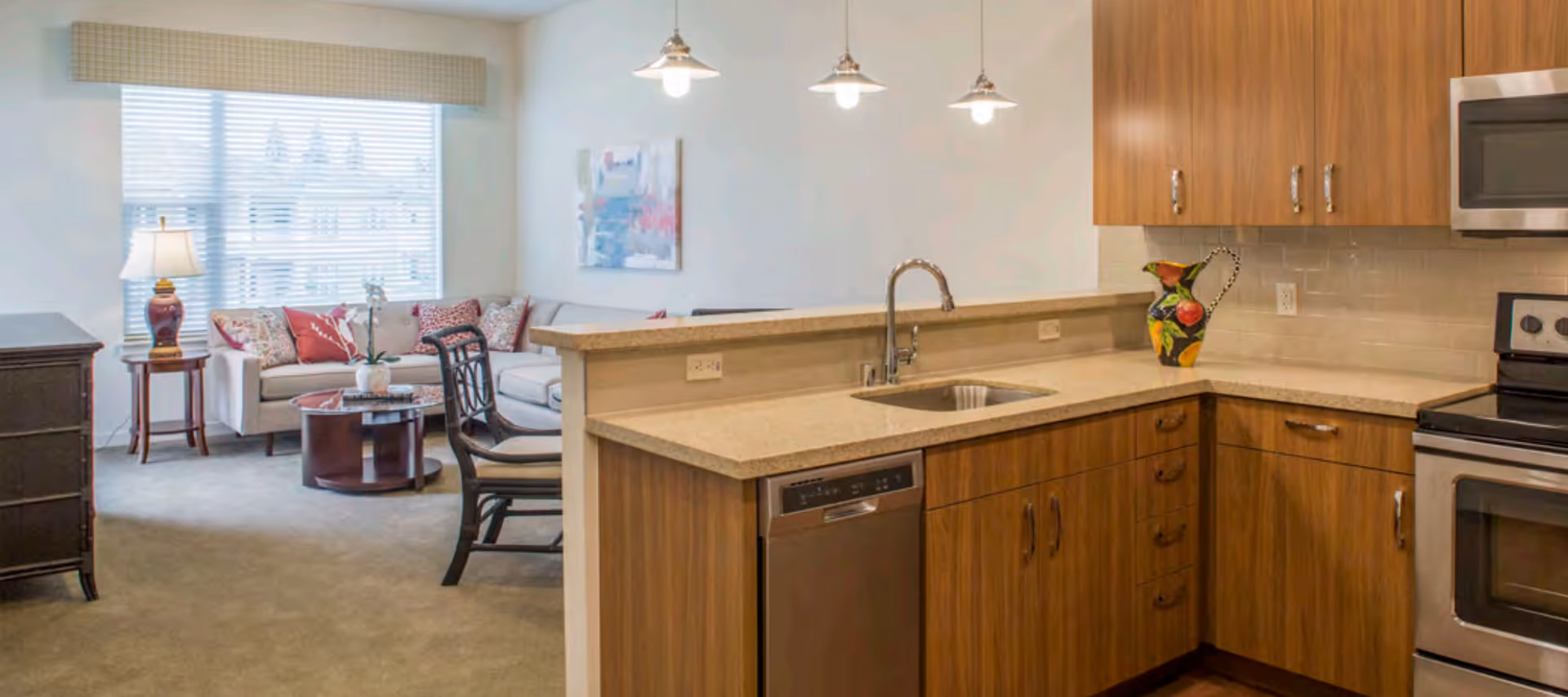 A modern open-concept living space featuring a kitchen with wooden cabinets, a countertop with a sink, a dishwasher, and a stove. Adjacent to the kitchen is a living room area with a beige sectional sofa adorned with red and patterned pillows, a wooden coffee table, a side table with a lamp, and a large window with blinds allowing natural light to fill the room.