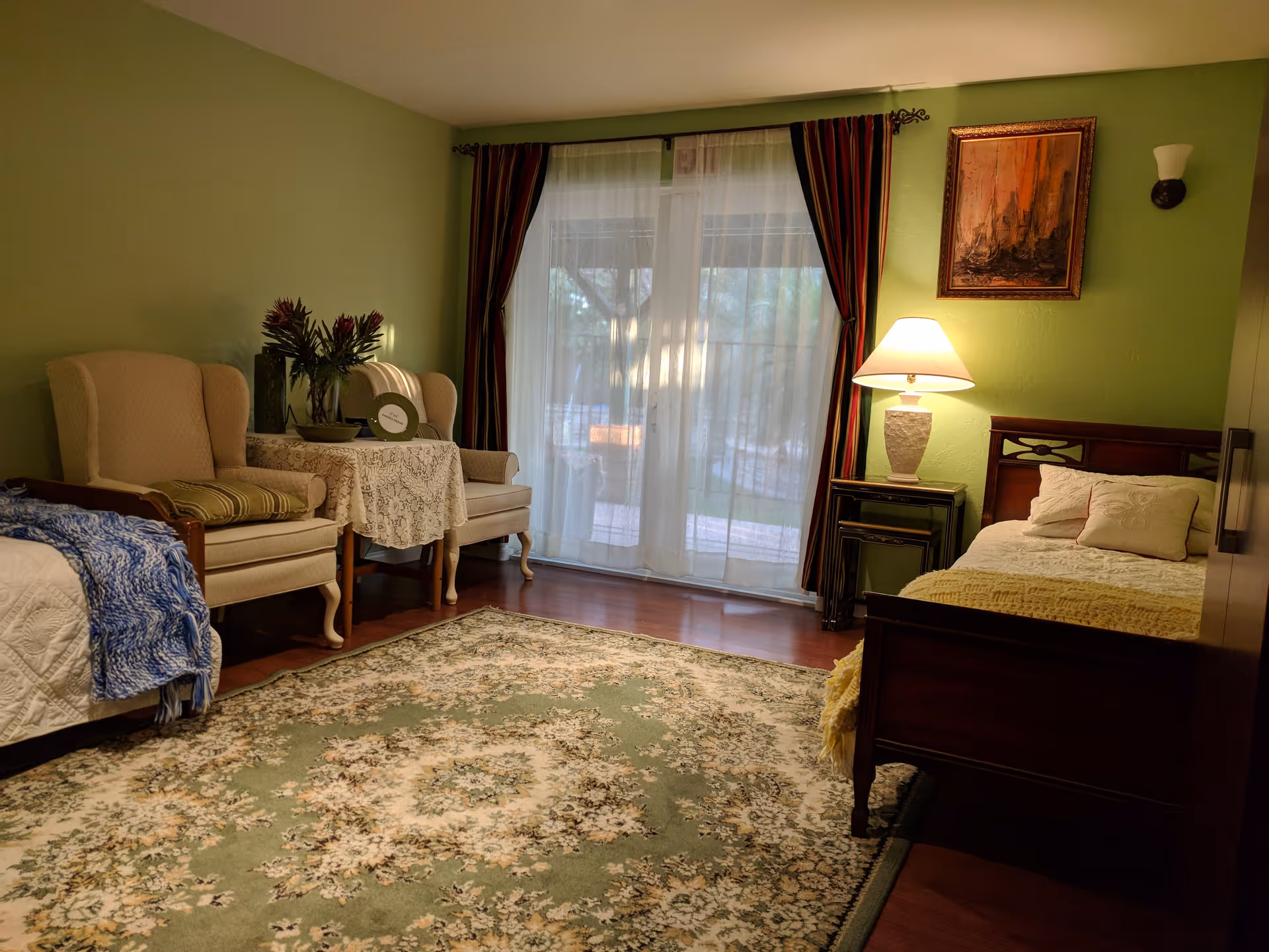 A cozy bedroom with green walls, featuring a single wooden bed with pillows and a yellow blanket, a nightstand with a lamp, two beige armchairs with a small table covered by a lace cloth between them, a large patterned rug on the wooden floor, and a window with sheer white curtains and dark drapes letting in natural light.