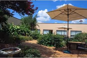 Courtyard with a patio table and umbrella surrounded by shrubs and a low building with windows under a blue sky.