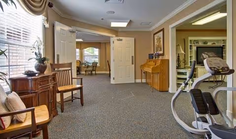 Interior view of a senior living facility hallway with carpeted floor, wooden chairs, a small wooden cabinet with a plant, a piano against the wall, and exercise equipment. The hallway leads to a room with large windows and seating visible in the background.