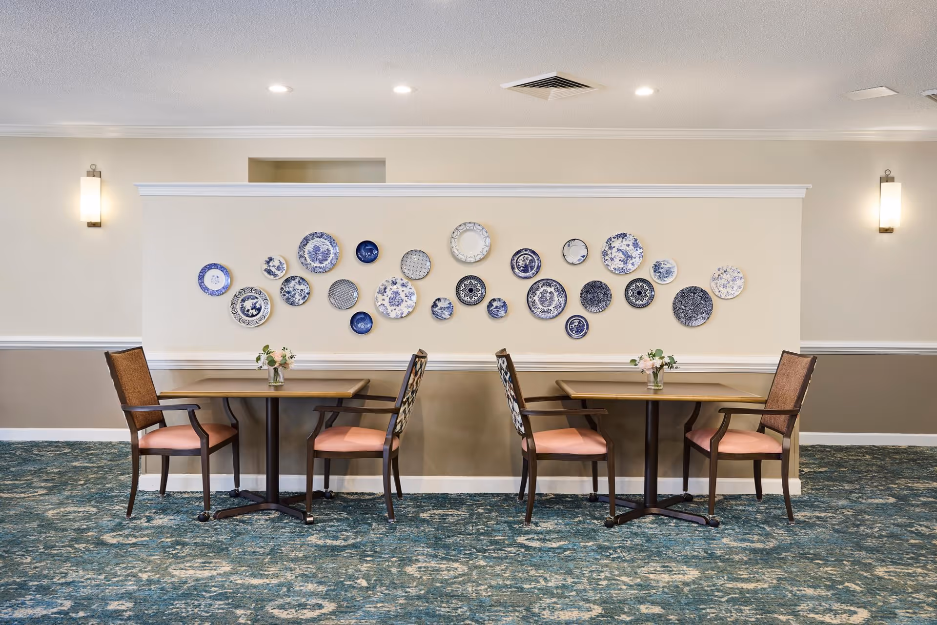 A dining area with two small square tables, each accompanied by two wooden chairs with pink cushions. The tables have small floral centerpieces. The wall behind the tables is decorated with a collection of blue and white decorative plates arranged in a horizontal pattern. The room has a blue patterned carpet and beige walls with white trim. Two wall-mounted light fixtures are visible on either side of the wall.