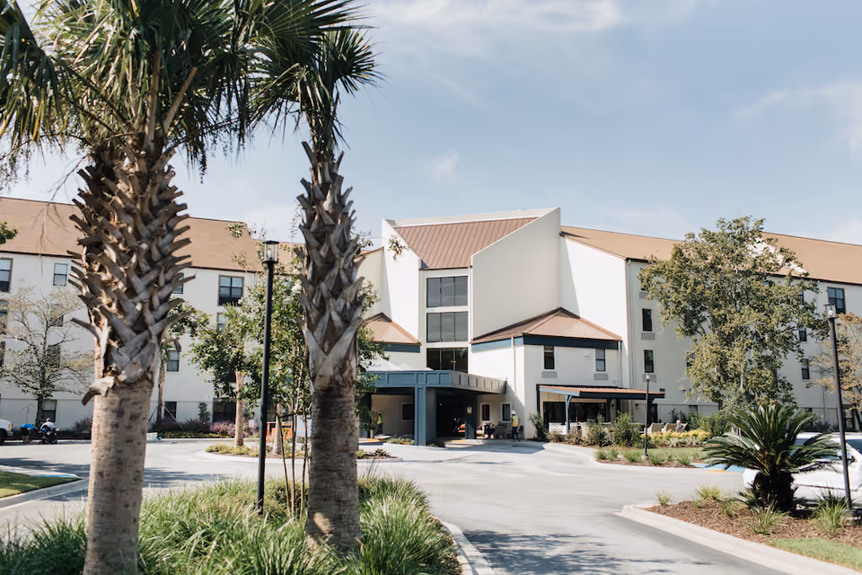 Exterior view of a senior living facility named The Social at Savannah, showing a multi-story building with a brown roof, white walls, and large windows. The entrance is covered by a canopy, and there are palm trees and landscaped greenery in the foreground under a clear sky.