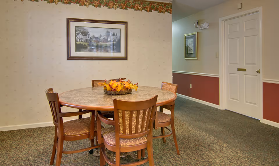 A small dining area with a round table surrounded by five wooden chairs with patterned cushions. A centerpiece with autumn leaves decorates the table. The walls have a beige wallpaper with a grapevine border near the ceiling. A framed picture of houses by a lake hangs on the wall. There is a white door and a hallway with another framed picture visible.