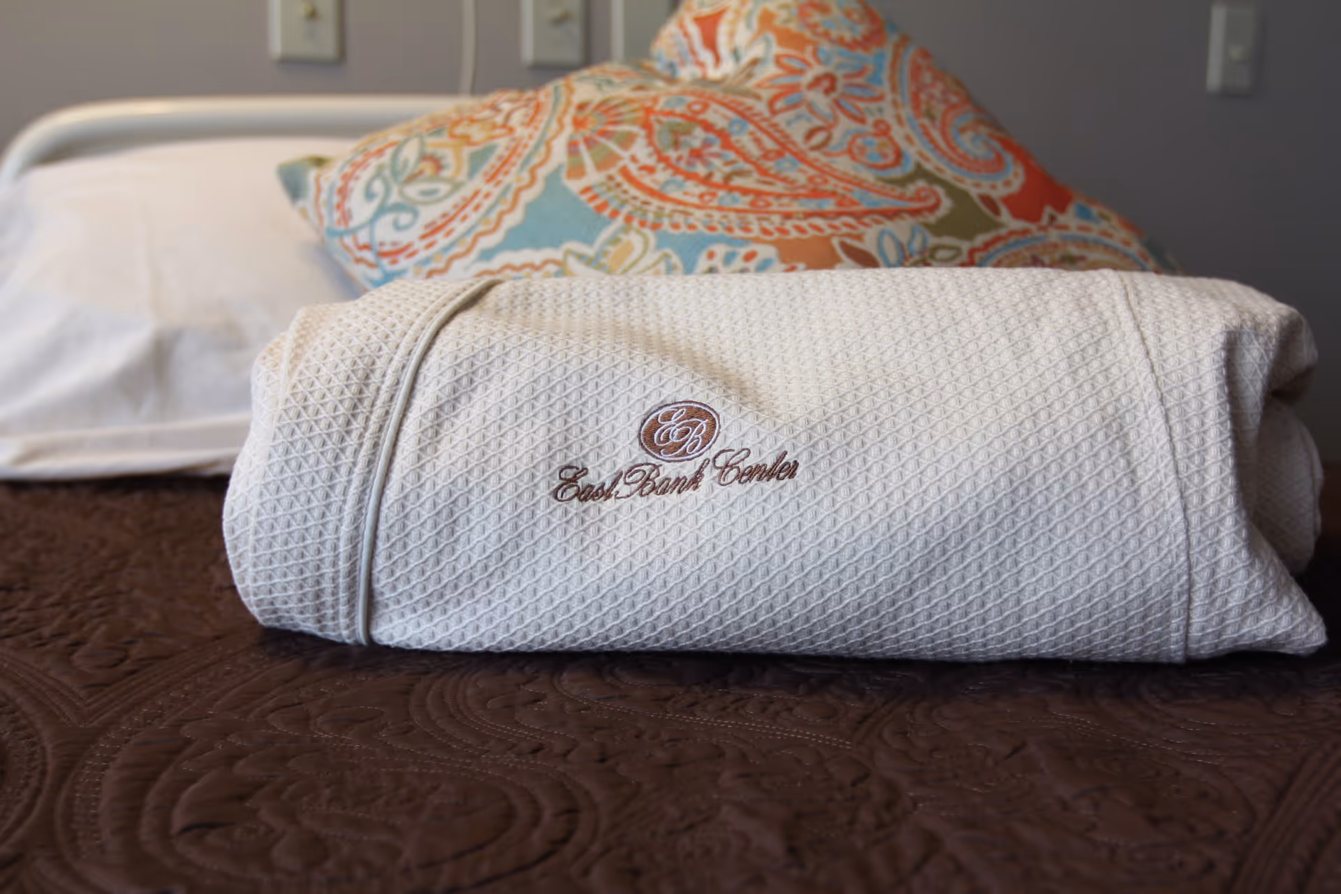 A neatly folded white textured blanket with the embroidered logo and text 'East Bank Center' placed on a brown quilted bedspread. In the background, there is a bed with a white pillow and a colorful patterned pillow against a gray wall.