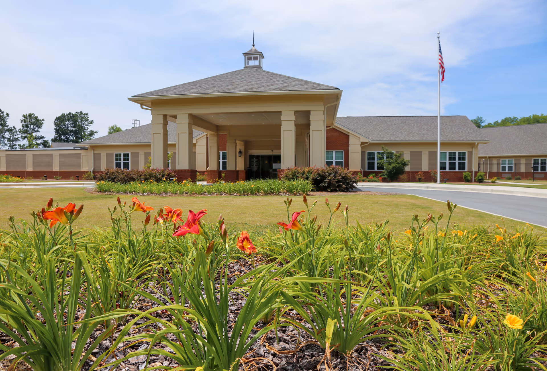 Front exterior view of Autumn Lane Health & Rehabilitation building with a covered entrance, a flagpole with an American flag, and a flower bed with orange and red flowers in the foreground under a partly cloudy sky.
