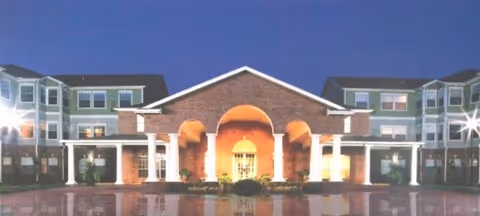 Front exterior view of a large senior living facility building at dusk with well-lit entrance featuring brick and white columns, multiple windows, and a clear sky background.
