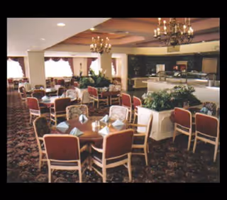 A spacious dining room with multiple round tables set with folded napkins and chairs around them. The room features chandeliers hanging from the ceiling, large windows with curtains allowing natural light, and decorative plants placed throughout. There is a buffet or serving area in the background.