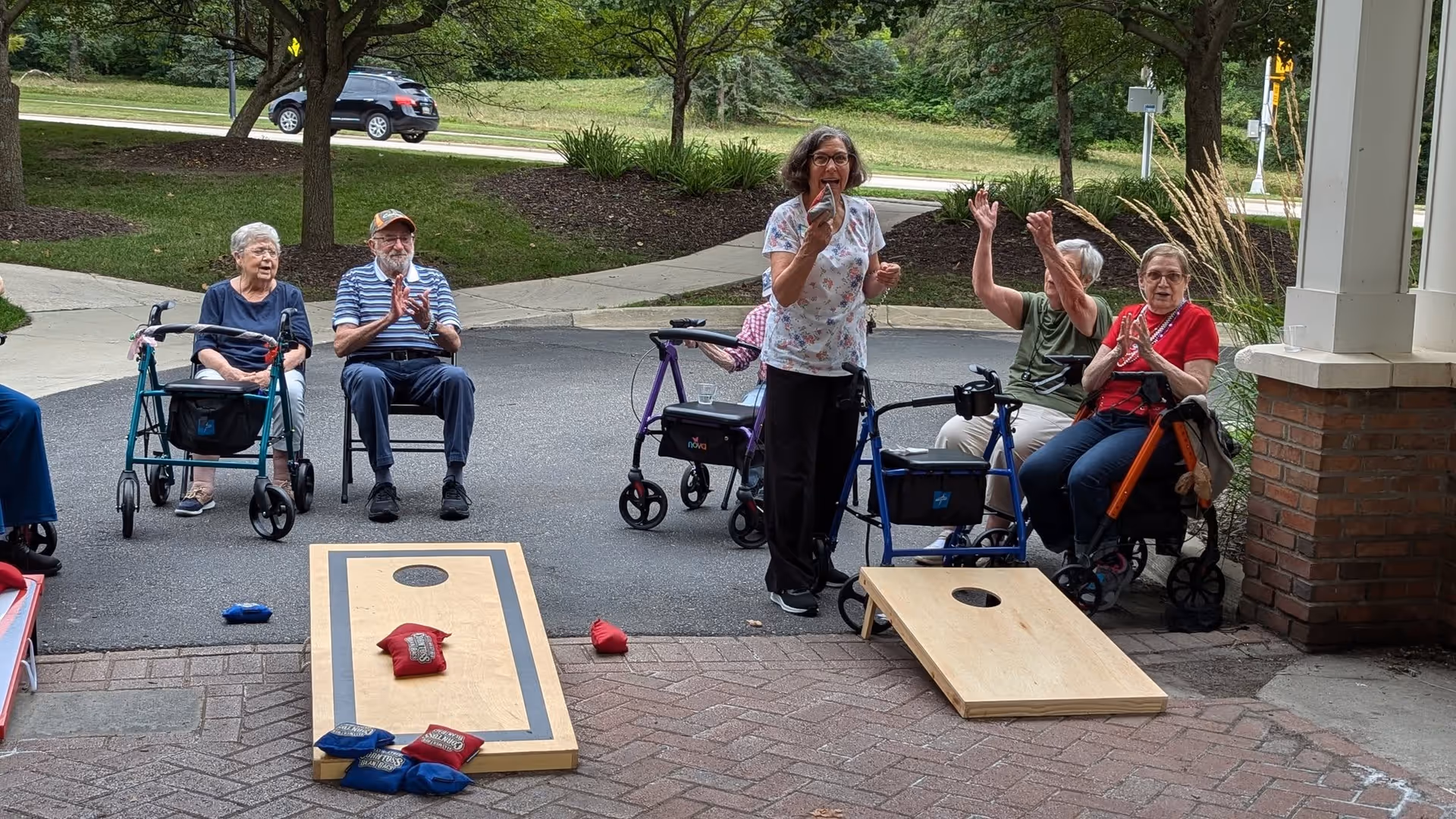 A group of elderly people sitting outdoors on walkers and chairs, playing a game of cornhole with bean bags on wooden boards. They appear to be enjoying the activity, with one woman standing and holding a bean bag, and others clapping and cheering.