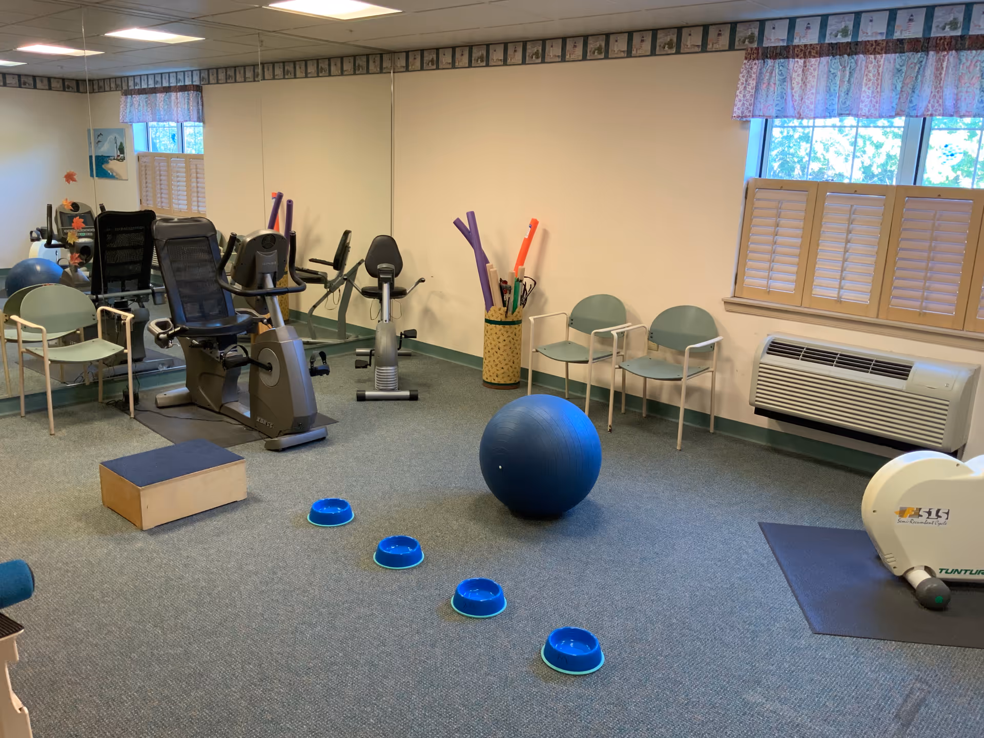 Small exercise room with stationary bikes, a large blue stability ball, balance bowls on the floor, chairs and windows.