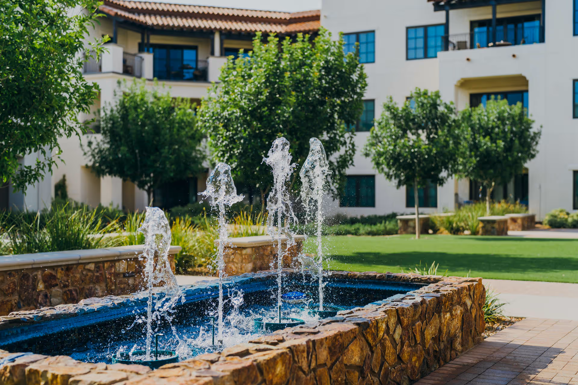 Outdoor courtyard area with a stone-bordered water fountain featuring multiple jets of water, surrounded by green grass, trees, and a multi-story building with balconies and large windows in the background.